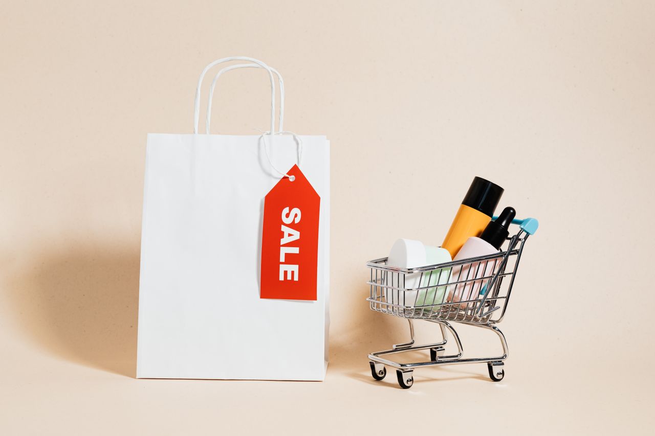 programs-01 White shopping bag with red sale tag and mini shopping cart on beige background.